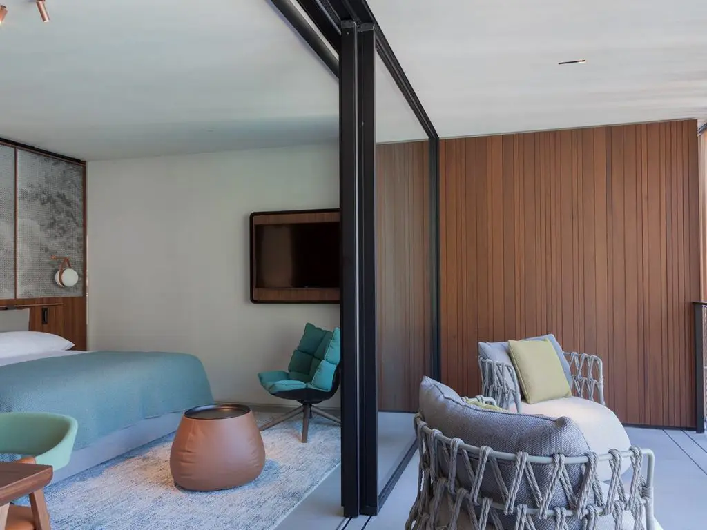 Hotel room with teal bed linens, green accent chairs and wood-panelled TV wall