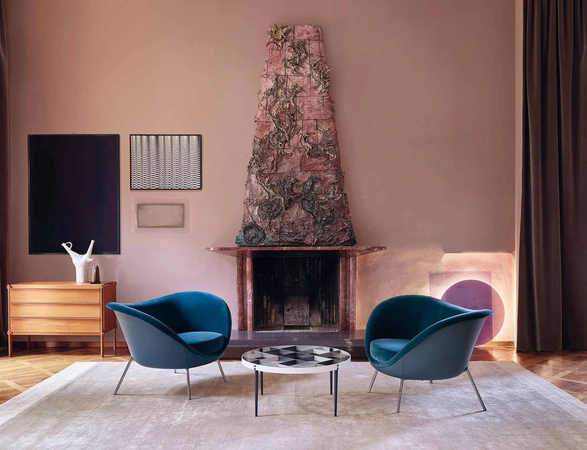 Living room with two teal velvet armchairs, a round coffee table and a tall decorative ceramic fireplace surround
