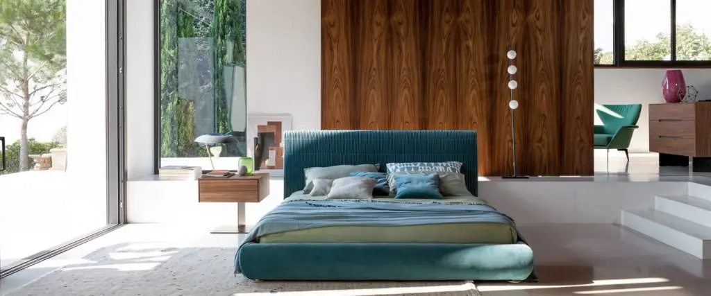 Bonaldo bedroom with a teal ribbed-headboard bed against a wood panel wall and floor-to-ceiling garden windows