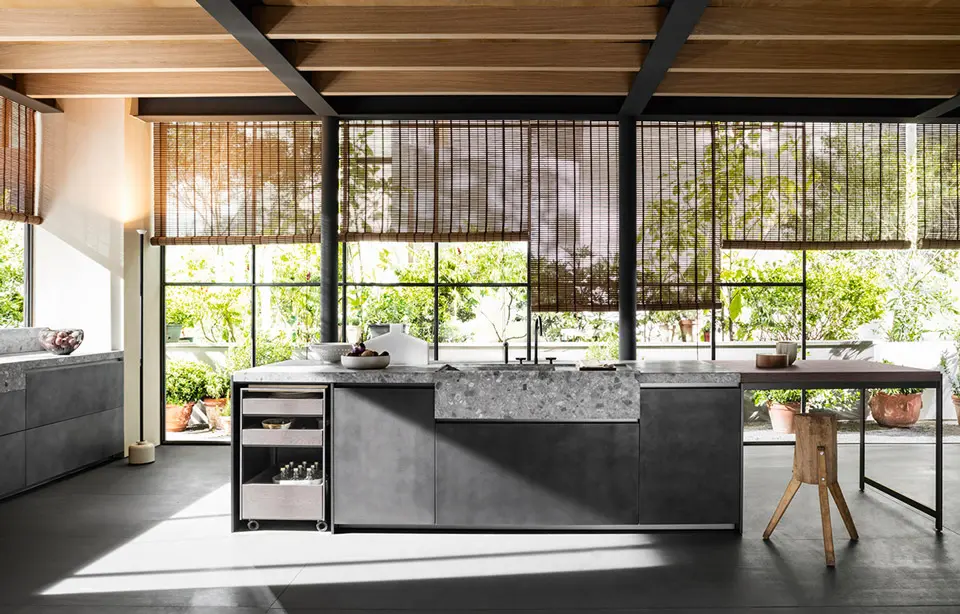 Grey stone kitchen island under wood ceiling beams with bamboo blinds and a garden view