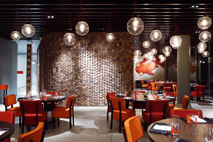 Restaurant with hanging glass globe lights, orange chairs and a round mosaic wood wall