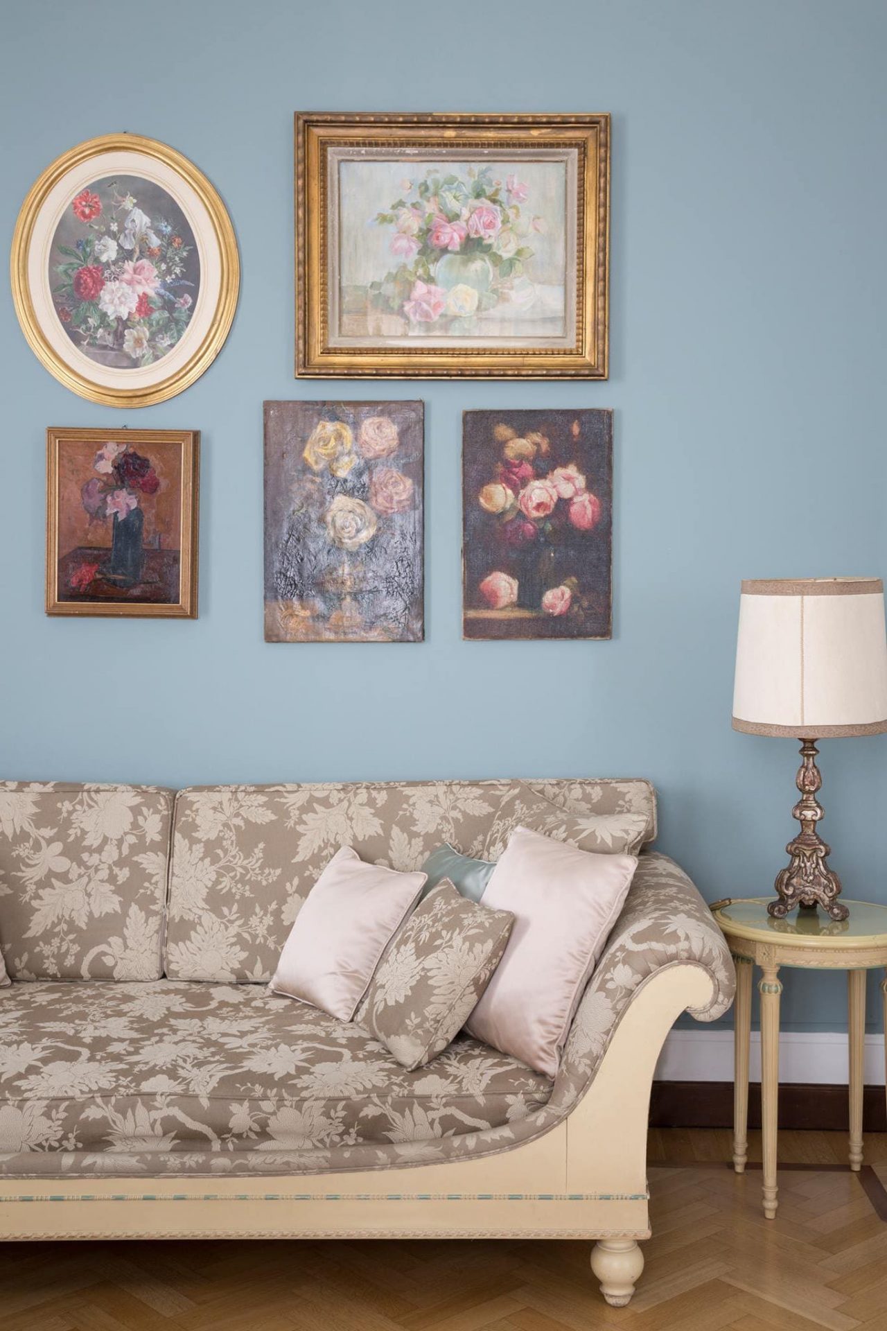 light blue wall with five framed art pieces, a lamp on a table and part of a couch with three pillows