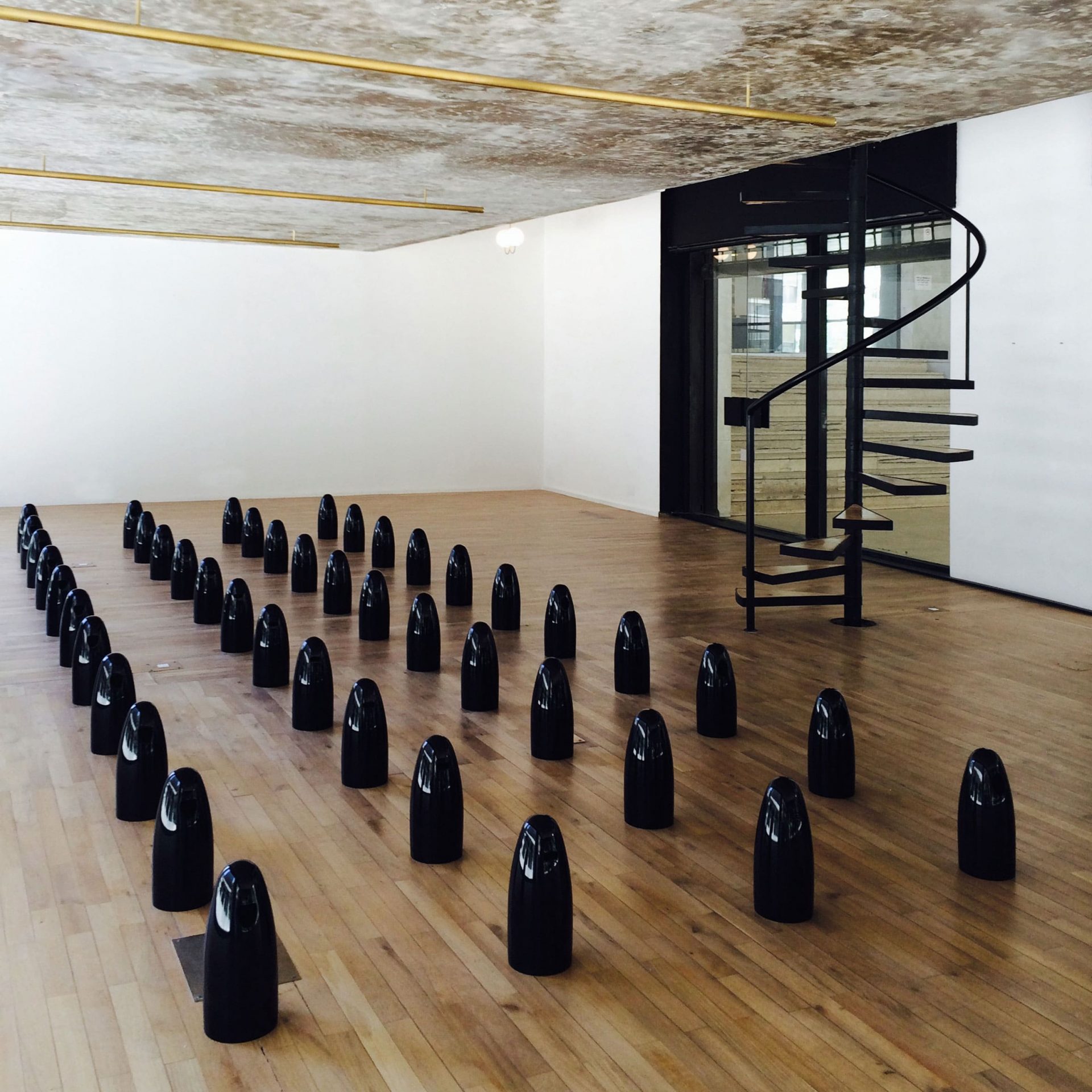 small black ceramic dolls are lined along the wooden floor of a small room with white walls and a spiral staircase