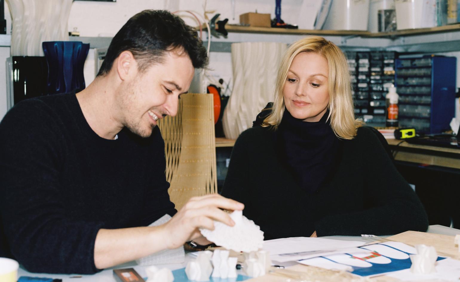 a man in a black shirt with black hair sitting at a table with a woman with blonde hair in a black shirt working in a studio