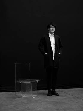 man with black hair wearing a suit standing up next to a glass furniture