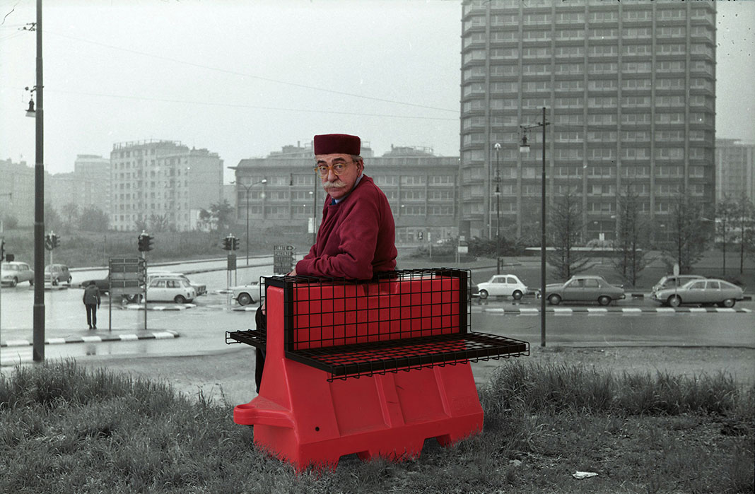 old man with black hat sits on a red bench overlooking a black and white cityscape